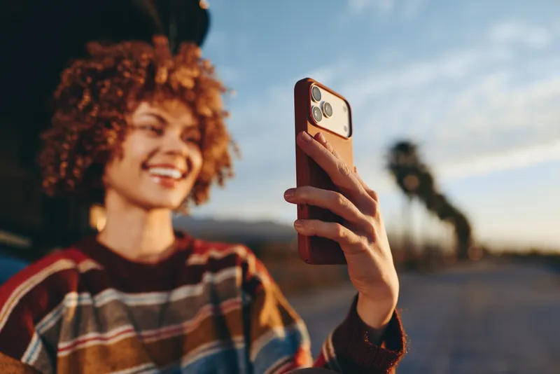 Woman outdoors smiling for a selfie with a smartphone, curly hair glowing in sunset light, casual lifestyle portrait, relaxed hands gesture, blurred street background