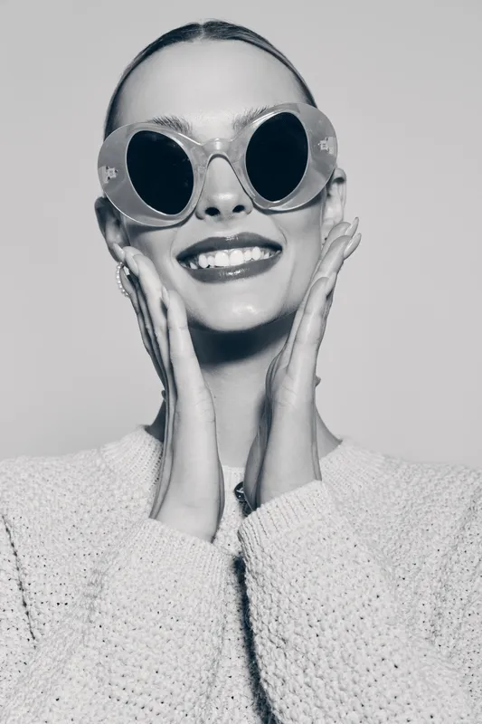 Smiling caucasian woman with fair skin wearing large round sunglasses and knitted sweater touches her face. Studio portrait with joyful mood and fashion concept in black and white.
