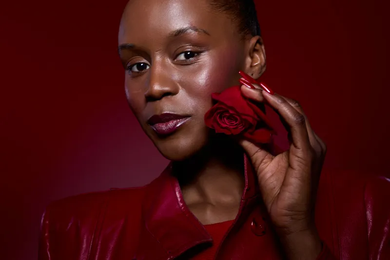 Close-up of dark skinned African woman holding red rose near face wearing red leather jacket. Studio portrait with red background. Concept of beauty, elegance, and natural skin glow.