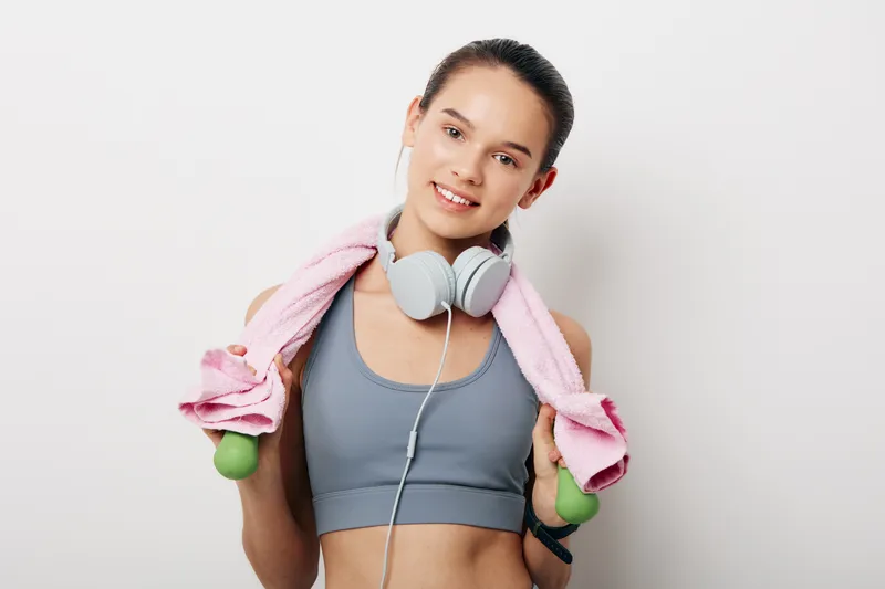 Active woman in gym wear after workout, with headphones around neck and towels over shoulders, holding light dumbbells, ready for a fitness routine and motivation in a bright setting.