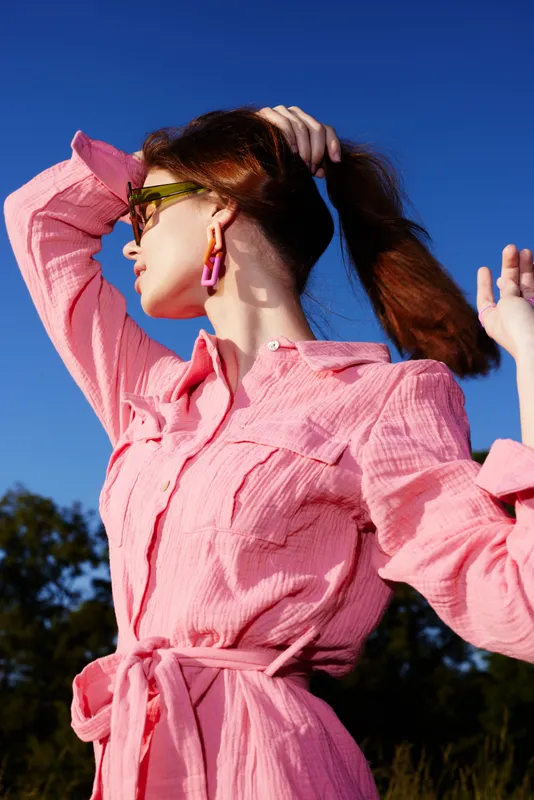 Young woman in pink dress posing confidently against a clear blue sky, showcasing modern fashion and style with vibrant colors and playful accessories