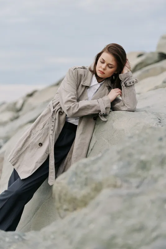Young woman in beige coat leans on large rocks near the coast. Overcast sky and soft natural light create calm mood. Concept for fashion, autumn, and thoughtful moments outdoors.