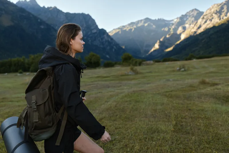A serene female hiker traverses a grassy valley toward rugged mountains. She wears a dark jacket and carries a large backpack with a rolled mat, enjoying expansive outdoor scenery.
