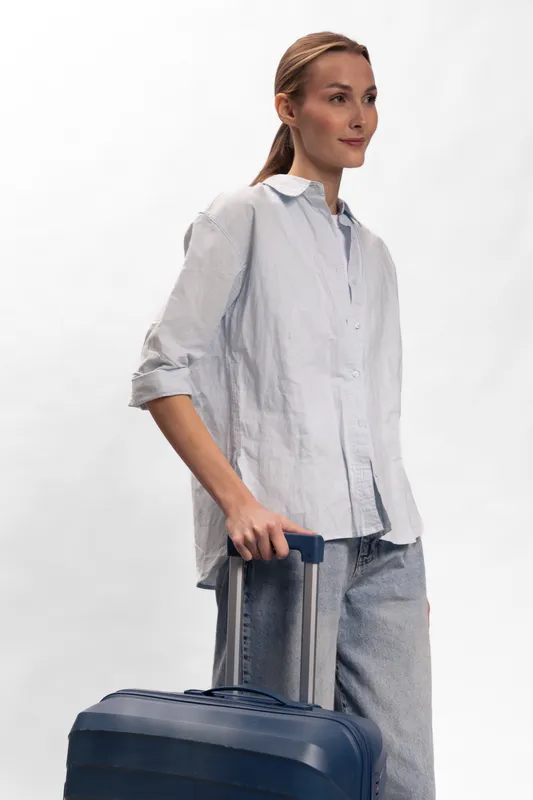 Young woman in casual clothes stands holding a suitcase handle, looking sideways with a neutral expression. Concept of travel, journey, and preparation for trip.