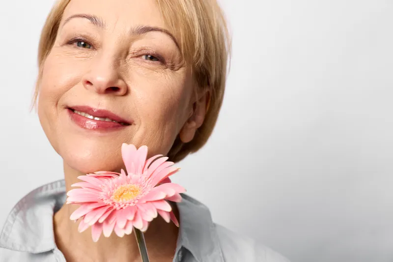 Woman, senior portrait indoors, holding a pink gerbera flower near her lips, gentle smile, soft light, calm eyes, neutral background, spring beauty mood, copy space, relaxed closeup