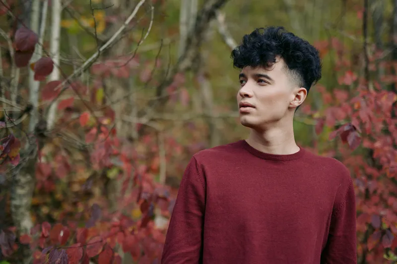 An authenticity driven portrait of a young man with curly hair in a maroon sweater, standing among autumn foliage, conveying credibility and genuine realism in a natural setting.