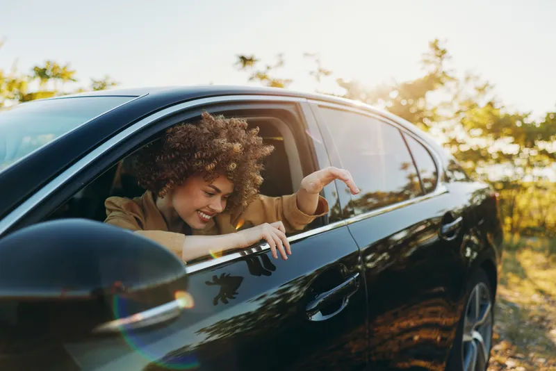 Smiling woman with curly hair leaning from a car window in a sunlit outdoor setting, capturing a candid lifestyle moment with retro film color and travel mood