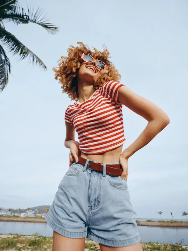 Confident young woman stands in a striped crop top, high waisted denim shorts, and belt beneath a bright blue sky, showcasing casual summer fashion and bold, playful attitude.