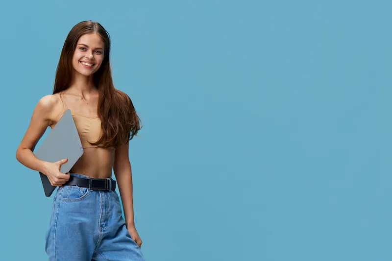 Young smiling woman holding laptop, wearing casual beige top and blue jeans, standing against plain light blue background with a relaxed confident pose.