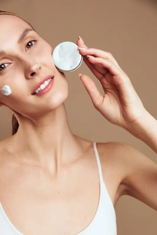 A young woman with clear skin applies white cream on her cheek while holding a round container in a bright studio. Concept for skincare, beauty, and facial care routines.