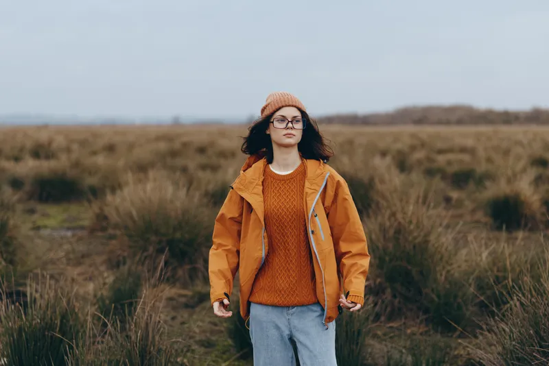 Woman lifestyle outdoors in a field, wearing a knit hat and orange sweater with a casual jacket, walking forward. Autumn nature background, soft daylight, relaxed portrait.