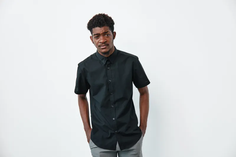 Young black man with dark skin wearing black shirt standing with hands in pockets against white background looking at camera.