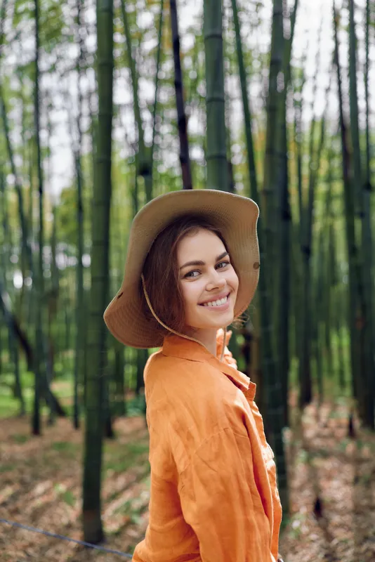 Woman smile hat bamboo forest portrait: young woman in orange jacket and wide brim hat smiling over shoulder in a serene bamboo grove, natural light and relaxed outdoor mood