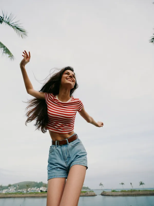 Young woman outdoors wearing a striped crop top and denim shorts raises arms joyfully under a bright sky, capturing a carefree moment of summer freedom and playful motion.