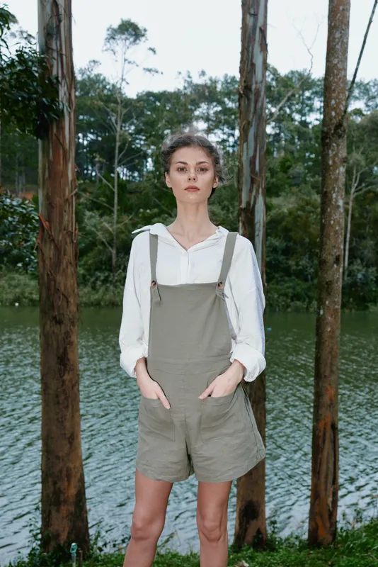 Elegant young woman in stylish khaki overalls standing by a serene lake surrounded by tall trees, showcasing a natural and relaxed vibe