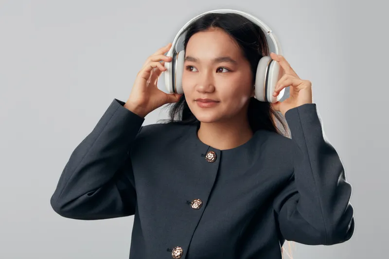 Young woman with headphones adjusting audio device on head looking away with smile isolated on gray background. Female person enjoying music or sound with casual dark jacket indoors.