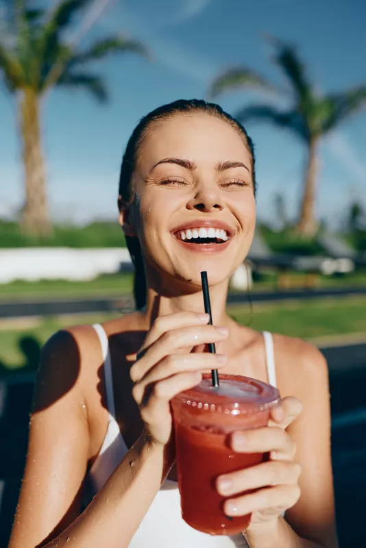 Smiling woman holding a cold red smoothie with a straw outdoors on a sunny day. Palm trees and clear sky in the background. Concept of lifestyle, health, and happiness.