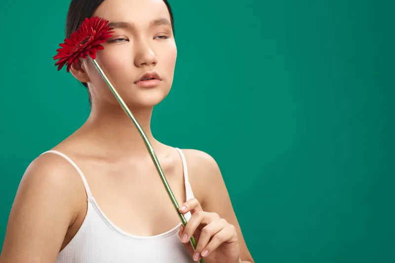 Asian woman softly holds a red flower near her face, eyes closed, posing against a teal backdrop, radiating serene beauty and minimal elegance in a stylized studio portrait.