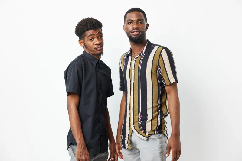 Two black men standing together, showcasing their unique styles and friendship, with an expressive, candid mood that highlights their personality and confidence.