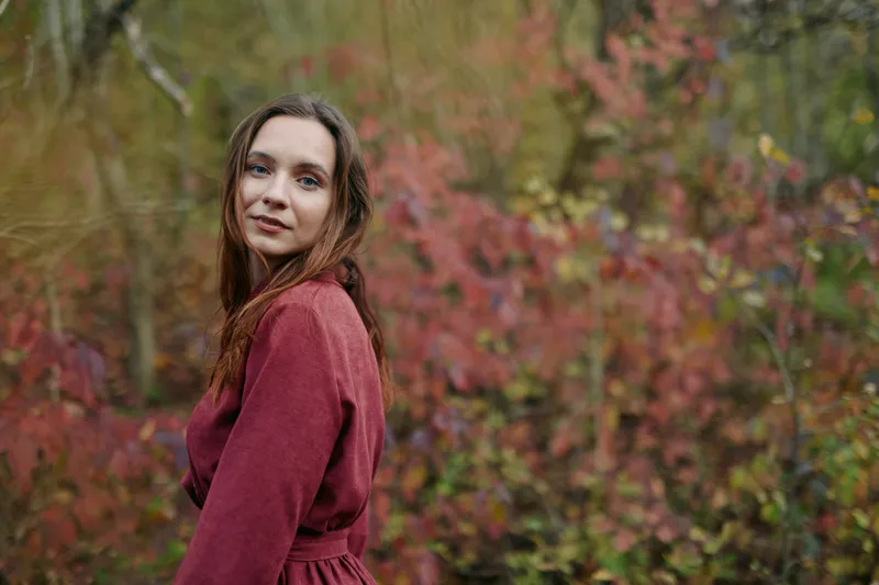 Portrait of a young woman in a simple burgundy dress standing among autumn foliage, natural pose and genuine expression conveying authentic, credible everyday emotion and warmth.