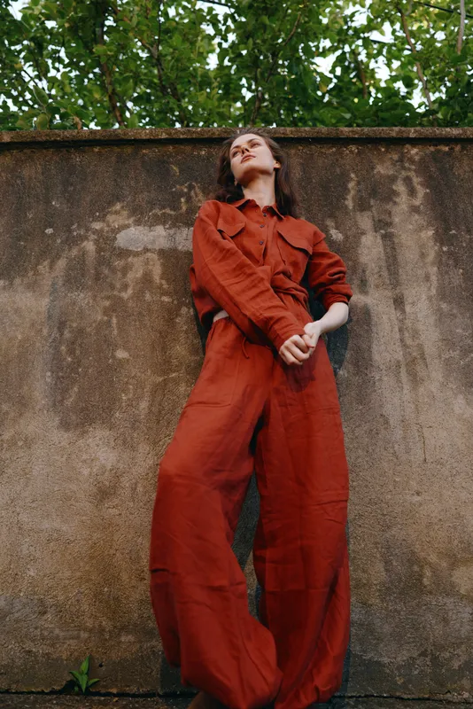 Woman in a red jumpsuit stands outdoors against a weathered wall, presenting a confident lifestyle moment, bold fashion statement, and lush greenery in the background