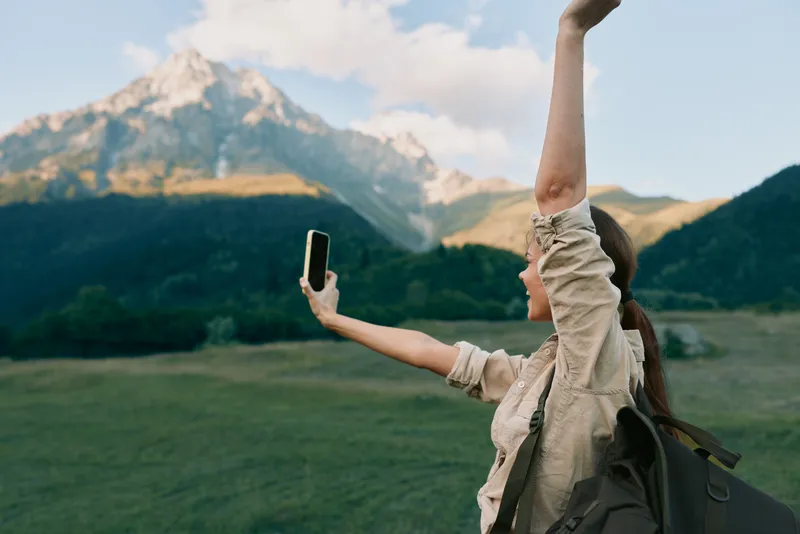 A woman outdoors takes a selfie with one arm raised, backpack on, against a mountain landscape. A lifestyle moment of exploration, freedom, and adventure in natural light.