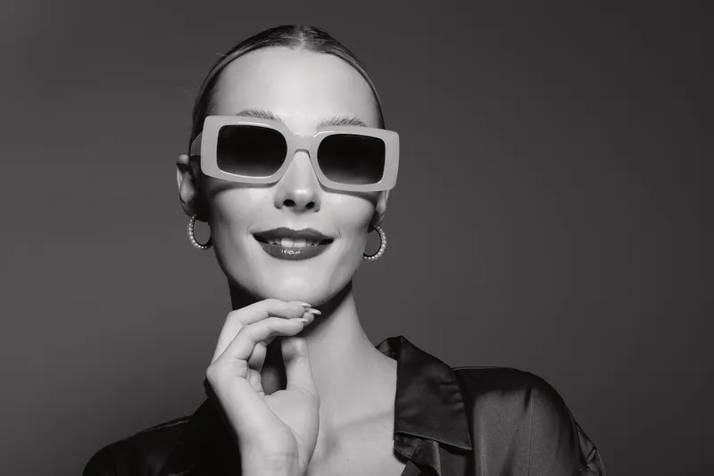 Young Caucasian woman with pale skin and slicked hair wearing large sunglasses and hoop earrings smiles confidently in black and white studio portrait fashion concept.