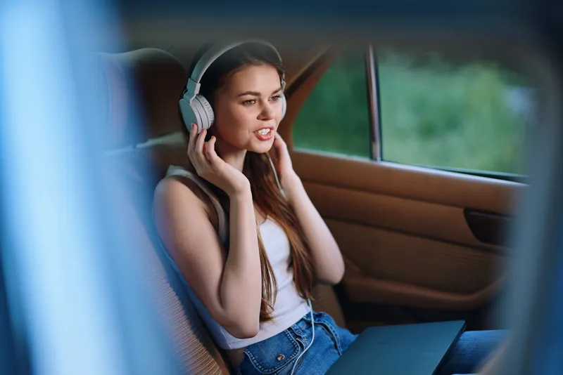 Young woman enjoying music in a car, wearing headphones, expressing joy with a laptop open, bright greenery outside, vibrant colors, summer vibes, relaxation.