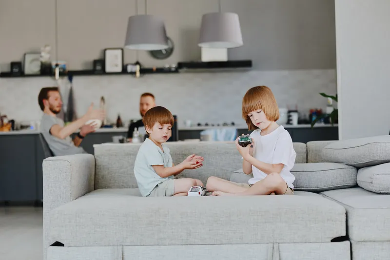Two children playing together on a sofa in a modern living room, while a man is interacting in the background. A cozy family atmosphere is present, fostering connection.