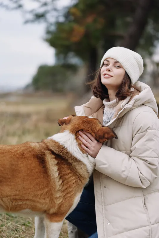 Young woman in a warm coat and beanie cuddles a friendly dog outdoors in a wide field, enjoying crisp air, gentle autumn light, and peaceful countryside companionship.