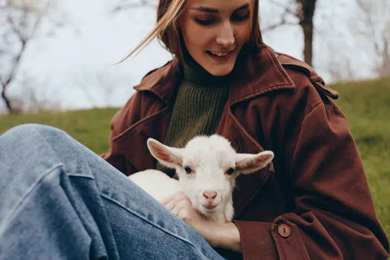 Smiling woman in a brown jacket sits outdoors with a kid goat, showcasing countryside lifestyle, companionship, and gentle care. Soft natural lighting enhances warm mood and well being.