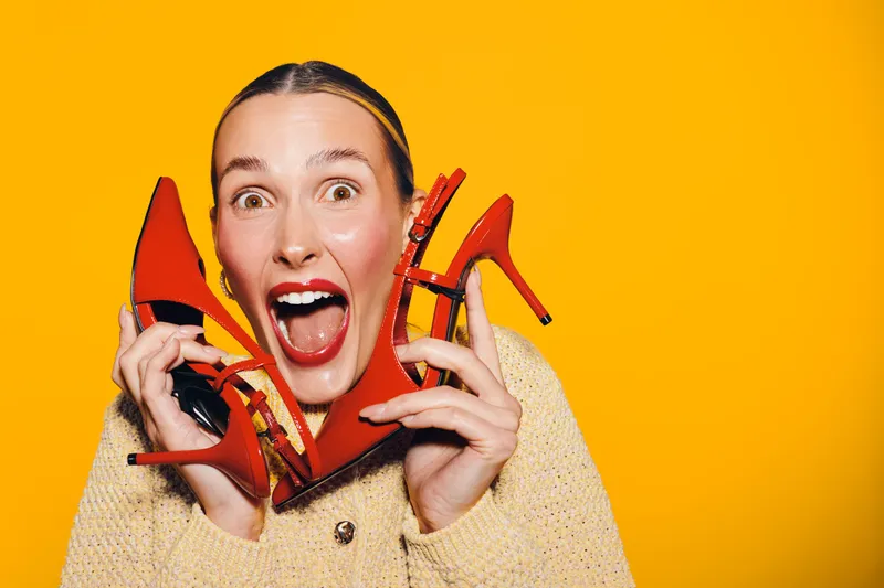Excited woman holds red high heels near face with wide open mouth on yellow background. Concept of fashion, shoes, shopping, happiness, and vibrant style in studio lighting.