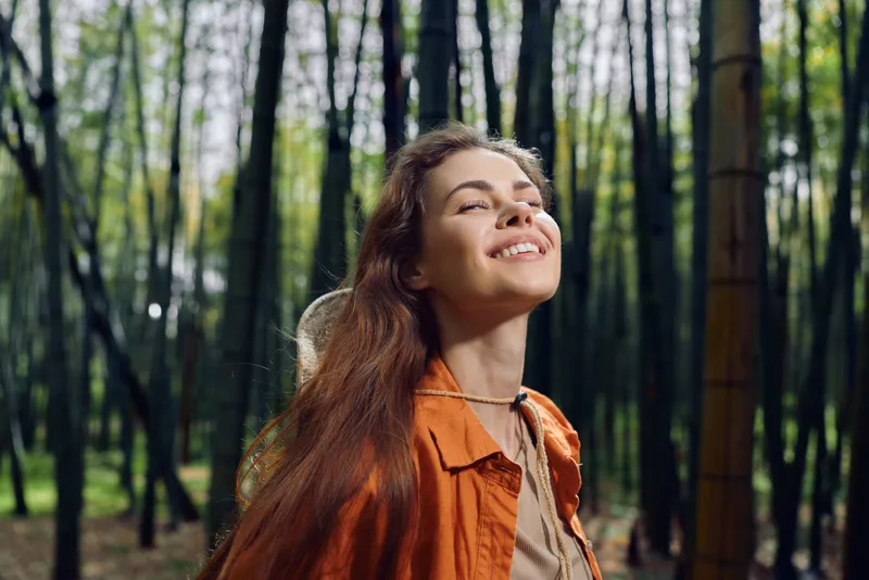 woman portrait smile in bamboo forest, nature outdoor scene with happy traveler enjoying sunlight and relaxation. Closeup of joyful face, long hair, casual jacket and peaceful escape