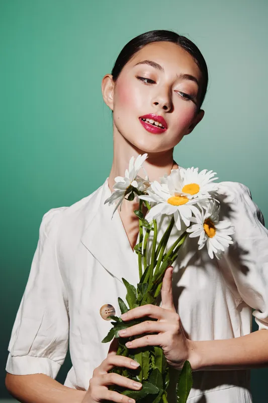 Fashion model in elegant white dress holds fresh daisies, looking down with soft expression. Creative beauty shot with green background and natural makeup, conceptual style.