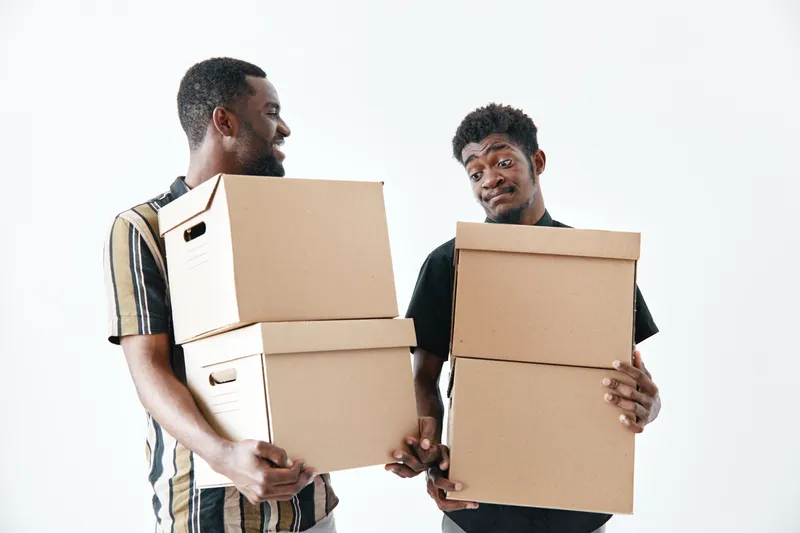 Two african american men carrying cardboard boxes for moving or delivery, teamwork and cooperation with casual clothes on white background, young black men holding packages together.