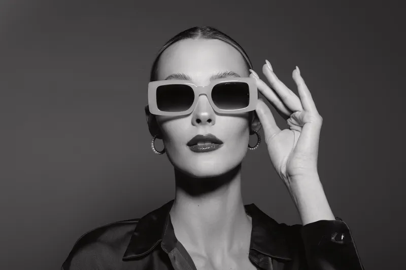 Stylish young Caucasian woman with pale skin wearing oversized sunglasses and hoop earrings poses confidently in black shirt. Monochrome portrait with fashion concept.