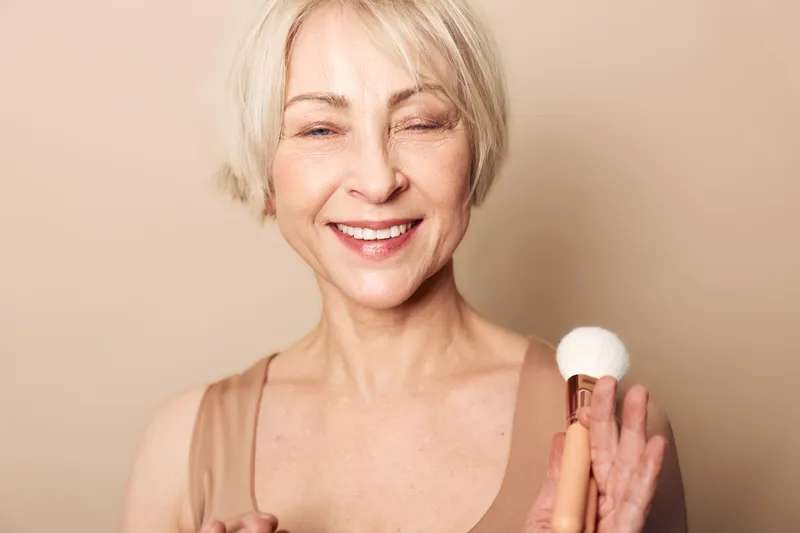 Smiling mature woman with short blonde hair holds makeup brush, applying powder on face with closed eyes. Concept of beauty routine, skincare, self-care, and natural makeup for older skin.