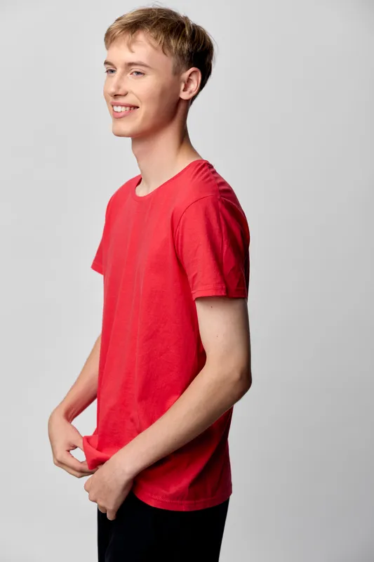 Young man in a red shirt with a bright smile, posing casually in a studio for fashion and lifestyle photography, showcasing youth energy and relaxed confidence.