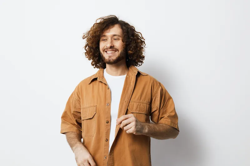 Smiling man with curly hair wearing a brown shirt and white t shirt, posing against a light background, ideal for lifestyle and personal growth themes