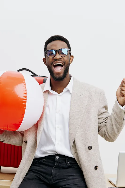 Cheerful smiling man with dark skin, wearing stylish sunglasses and light beige blazer, holding a beach ball over shoulder, relaxed and joyful mood in bright studio setting. People lifestyle concept