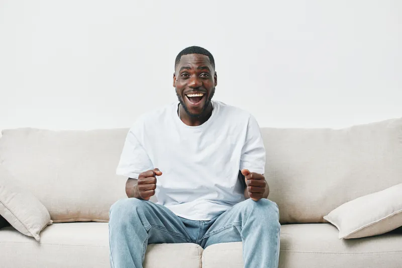 Happy young African American man sitting on a couch, celebrating success with excitement and joy, wearing a casual white t shirt and jeans