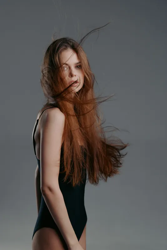 Young woman with long red hair wearing black bodysuit against gray background, delicate and contemplative mood