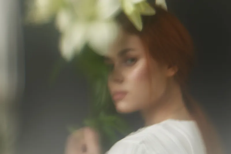 Beautiful woman with red hair and soft makeup, holding a bouquet of lilies in a serene indoor setting, showcasing natural beauty and tranquility
