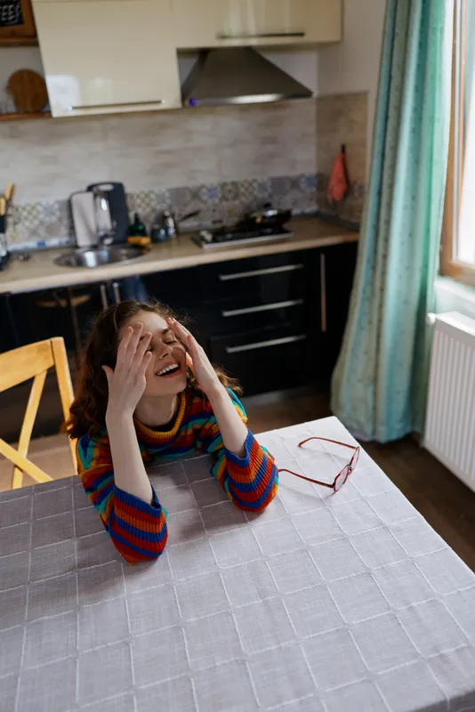 Young woman with a colorful striped sweater sits at a kitchen table laughing with her hands on her face, creating a vivid candid moment of cozy domestic joy indoors.