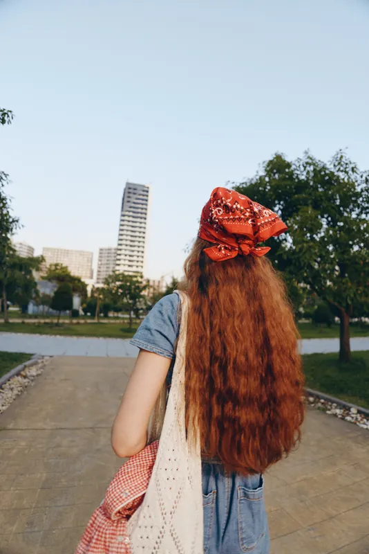 redhead woman with long hair walking in urban park wearing red headscarf and blue dress holding bag on sunny day in casual style