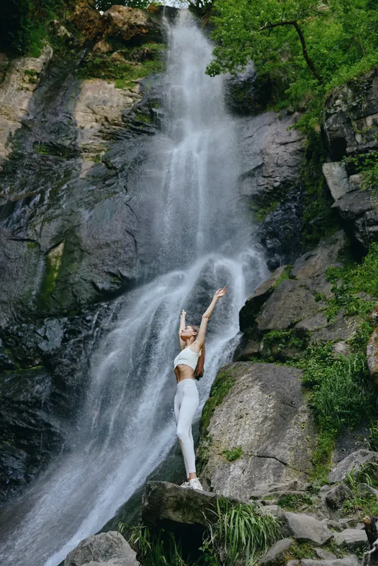 woman practicing yoga near waterfall in nature, wearing white activewear, serene atmosphere, surrounded by rocks and green foliage, outdoor fitness concept
