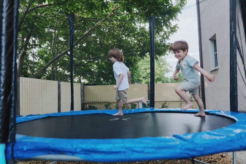 Two cheerful boys happily bouncing on a trampoline in a sunny garden, enjoying their playful day outdoors with joyful expressions and light clothing.