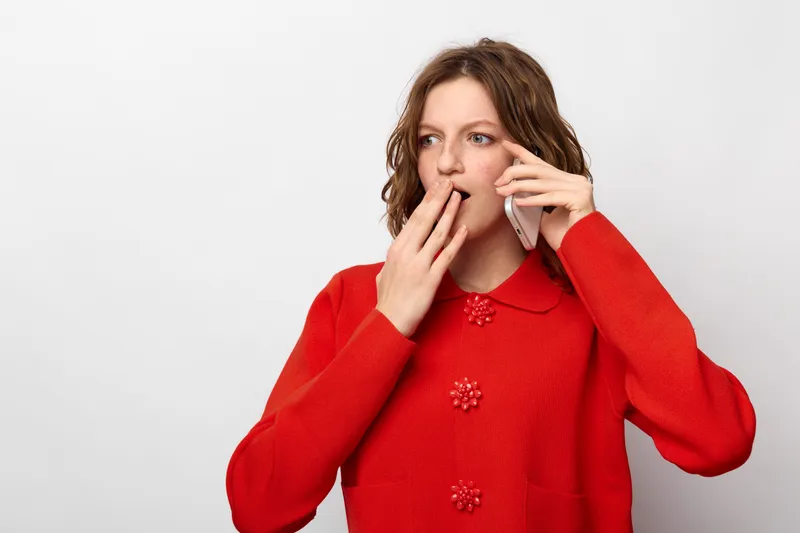 Surprised woman in red sweater talks on smartphone covering mouth with hand. Isolated on white background. Concept of shock, unexpected news, communication, and emotion.
