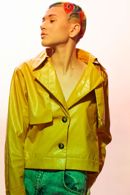 Young woman with colorful braided hair wearing a shiny yellow jacket and green pants, posing confidently against a soft pastel background in vibrant studio lighting.