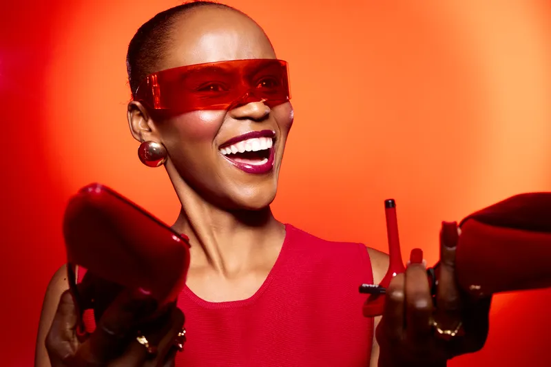 Happy African woman with dark skin wearing red dress and red sunglasses smiles brightly while holding red purse and smartphone on orange background. Concept of fashion, joy, and lifestyle.
